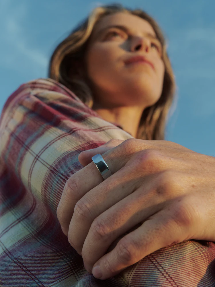 A person looking upward outdoors against a blue sky, wearing a plaid shirt while wearing an Oura Ring 5 in silver finish.