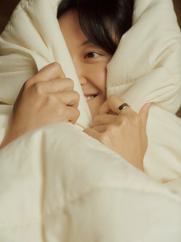 A smiling woman wrapped in a white comforter, wearing an Oura Ring 5 in gold finish.