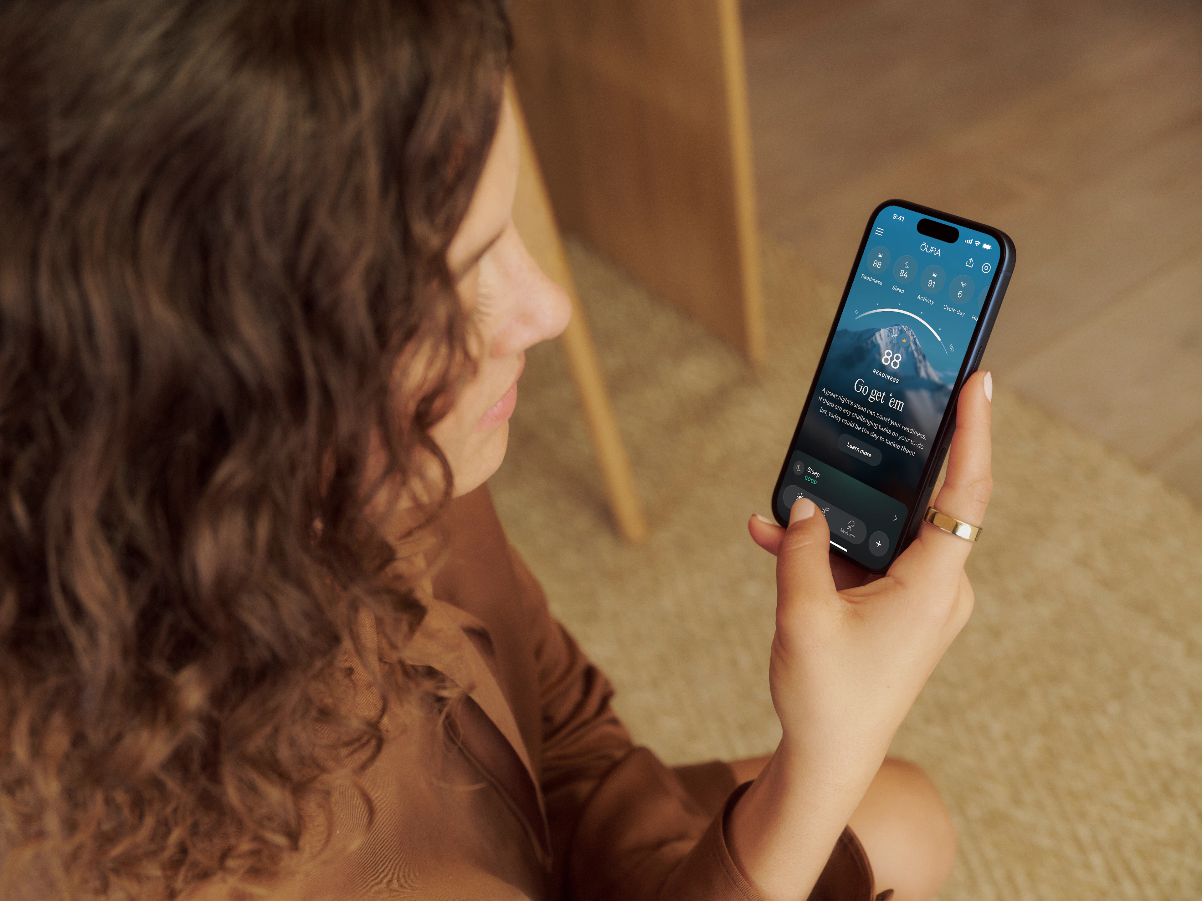 Oura Ring 5を着用した手でスマートフォンを持ち、Ouraアプリを見ているウェーブがかかった髪の女性。