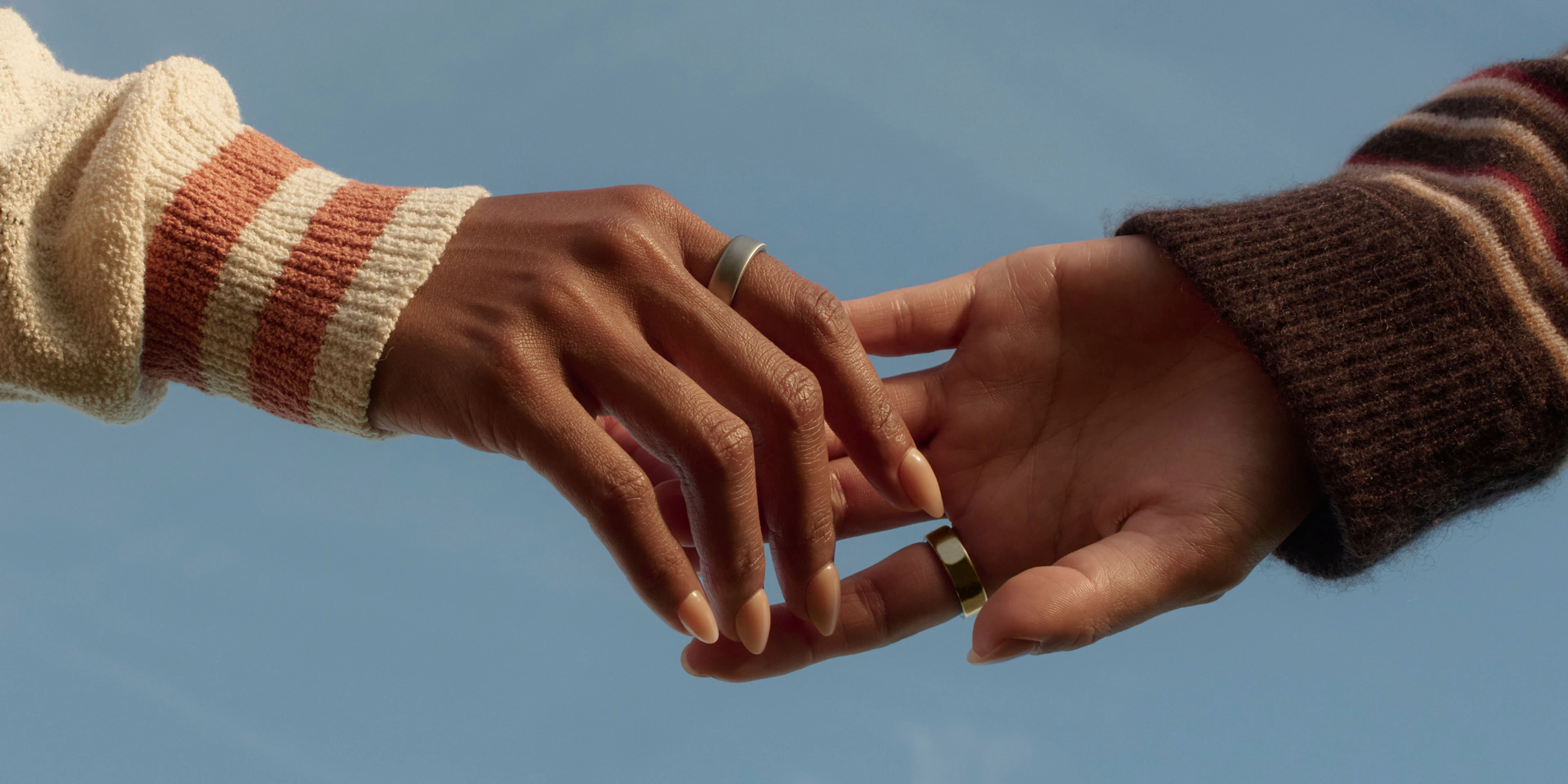 Two people's hands reaching toward each other against a blue sky, each wearing an Oura Ring.