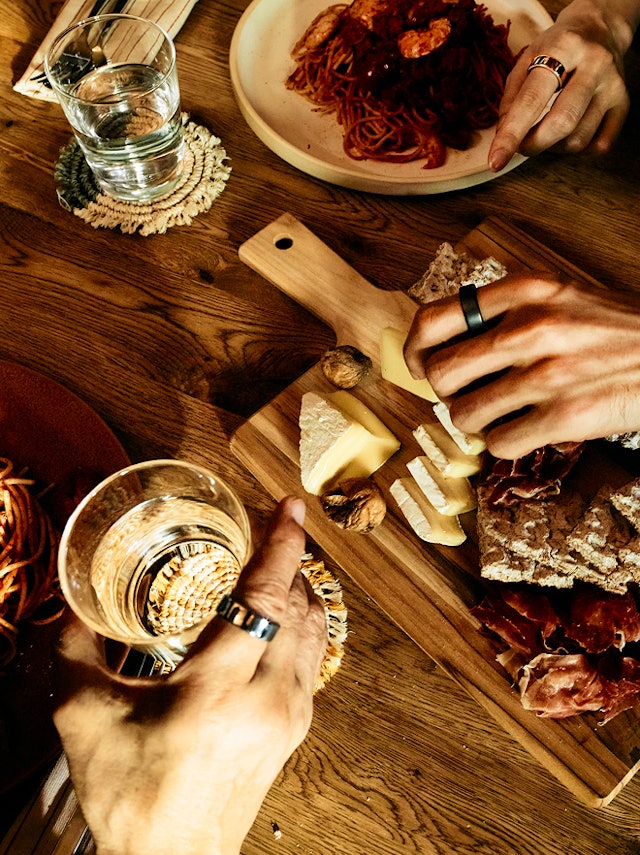 A close-up of three people's hands sharing a meal with pasta, cheese, and cured meats on a wooden table, all wearing Oura Rings