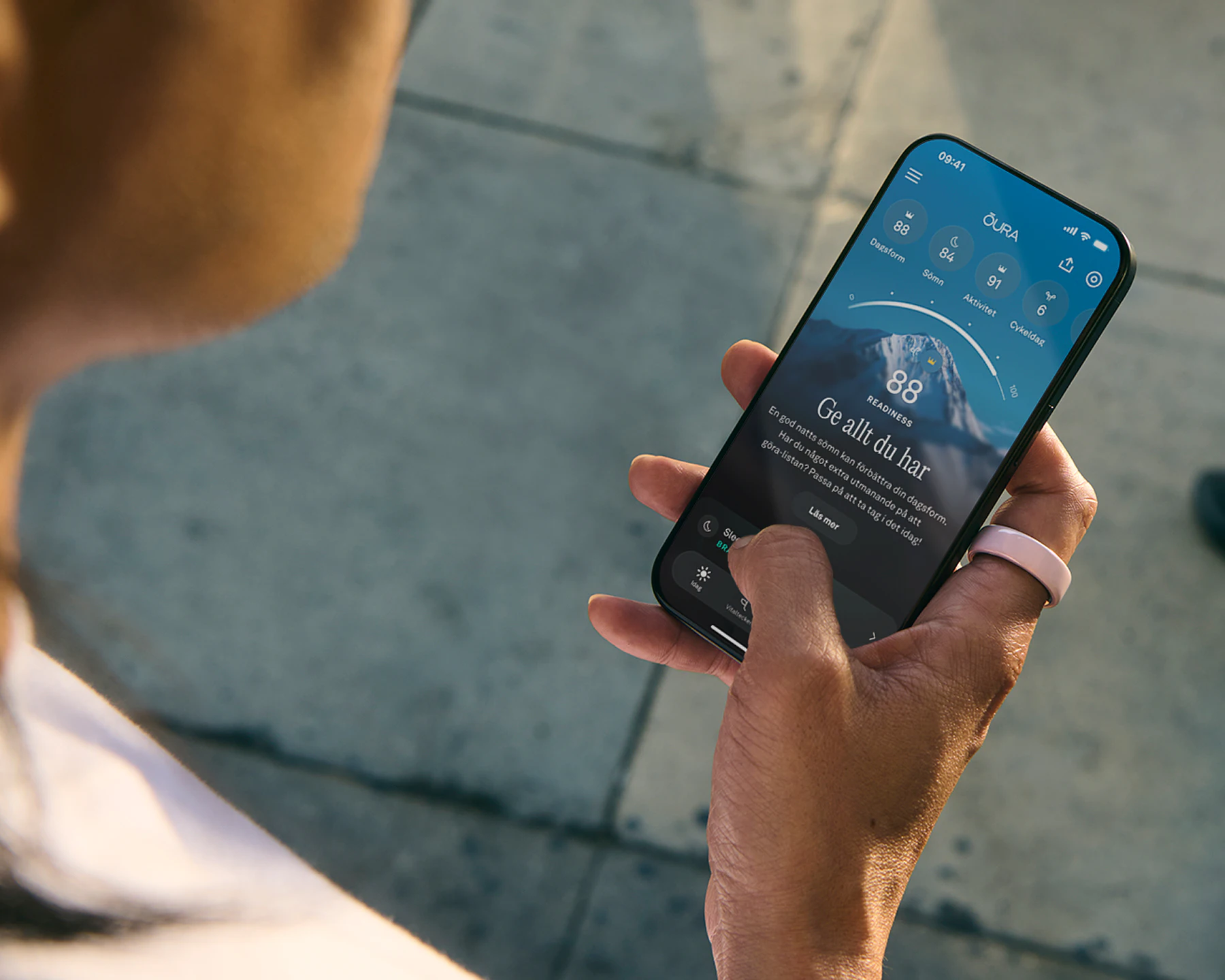 En person som tittar i Oura-appen på sin smarttelefon och bär en Oura Ring Ceramic i Petal-utförande på höger pekfinger
