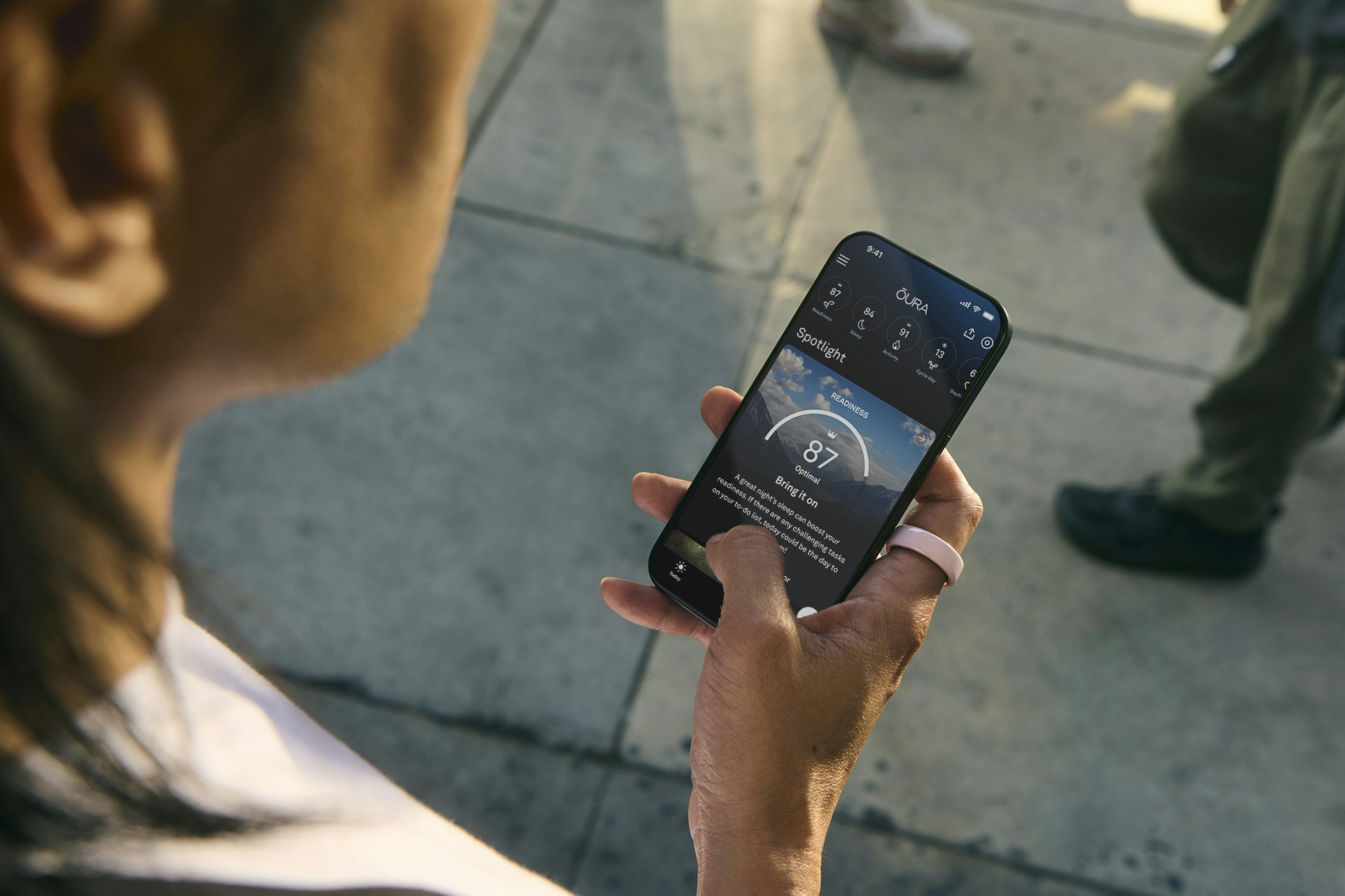 A person views the Oura app on their smartphone while wearing a Petal Oura Ring Ceramic on their right index finger