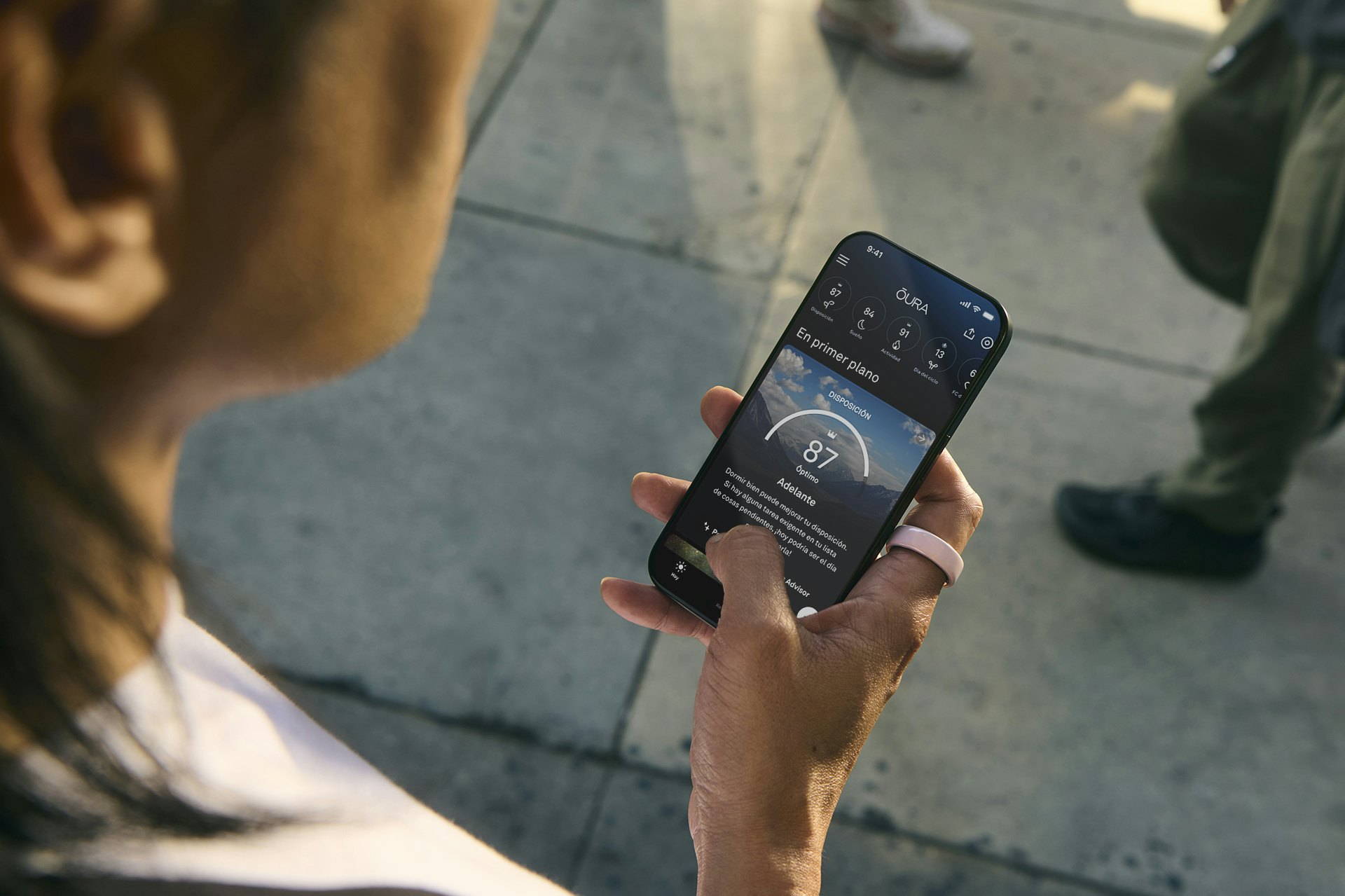 Una persona con un Oura Ring Ceramic con acabado Petal en el dedo índice derecho mira la aplicación Oura en su smartphone.