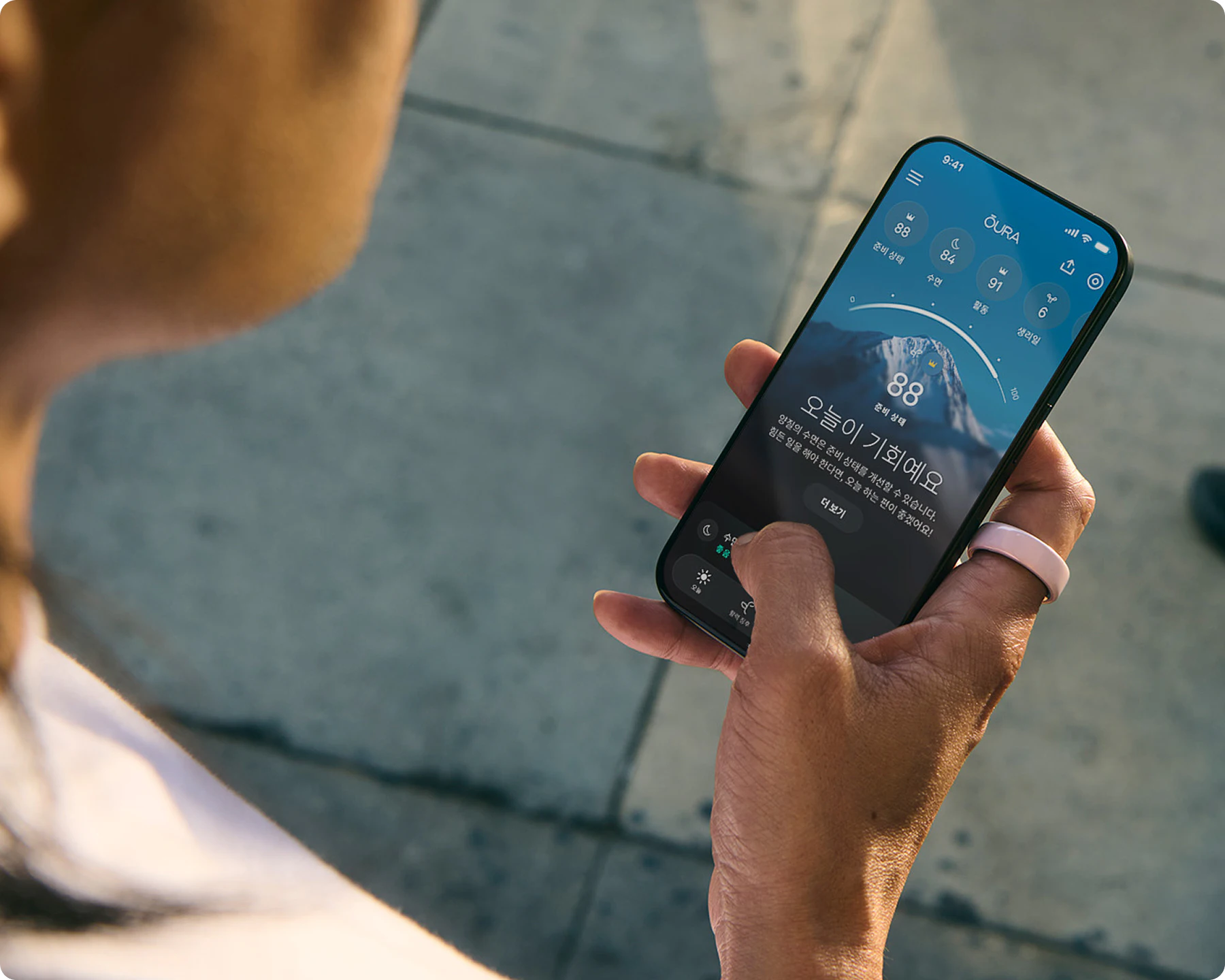 오른손 검지에 Petal Oura Ring Ceramic을 착용한 사람이 스마트폰에서 Oura 앱을 보고 있는 모습