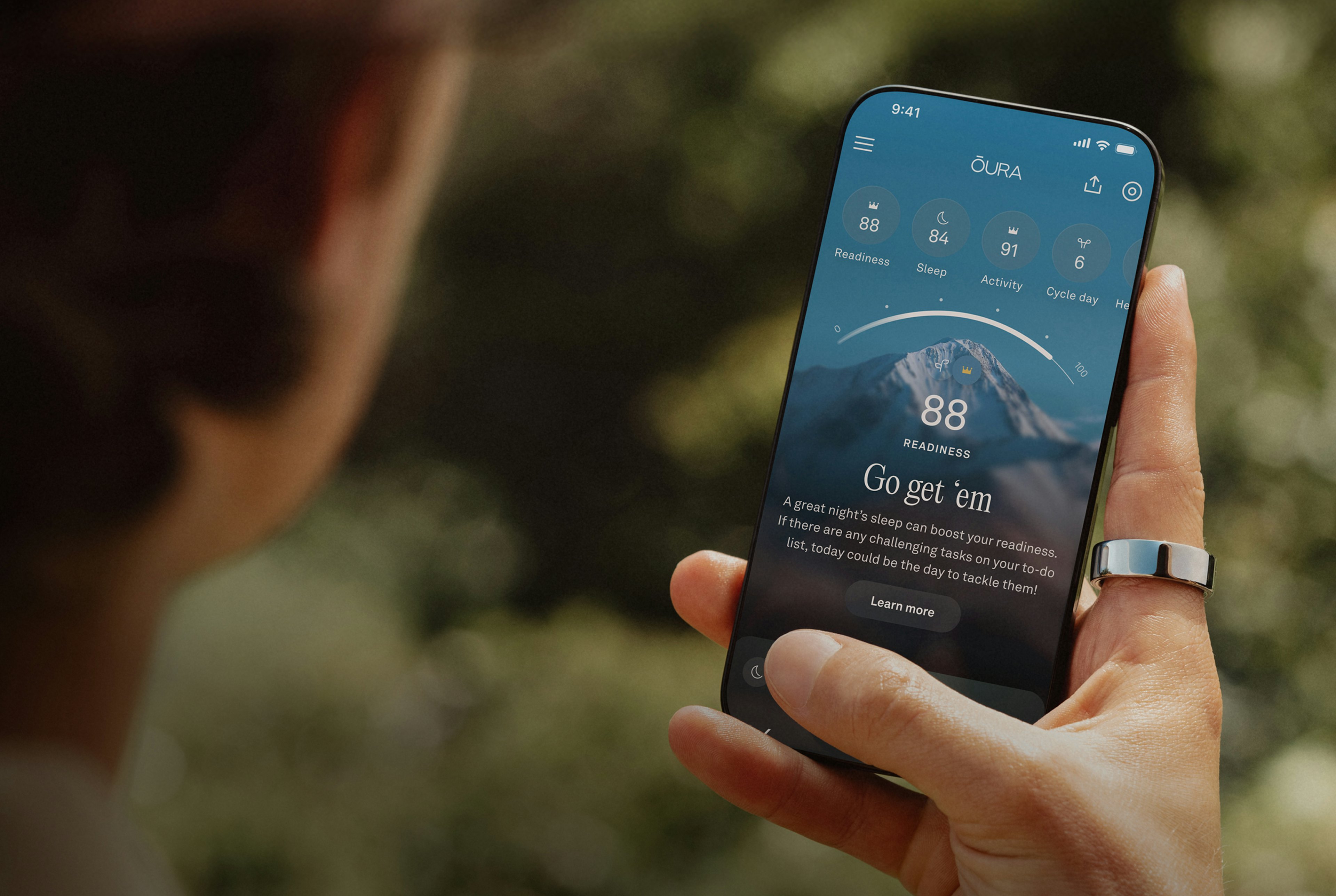 A person views the Oura app on their smartphone while wearing an Oura ring on their right index finger