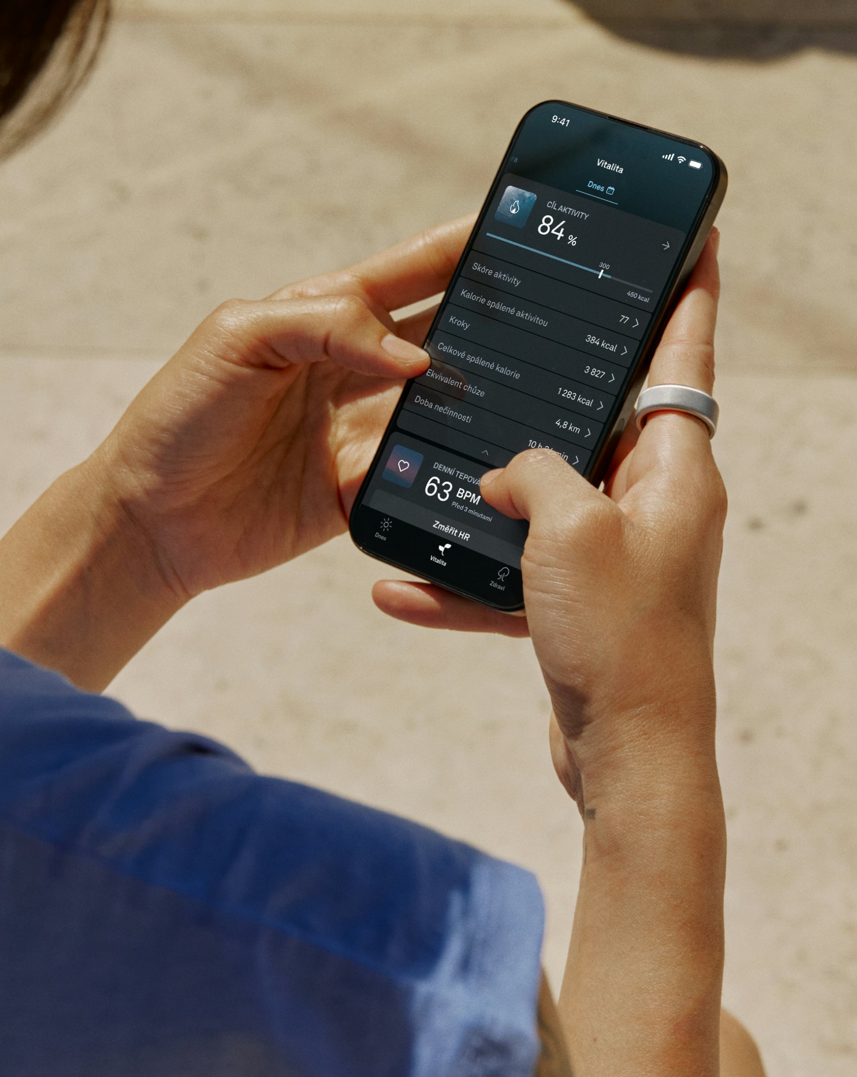 Händer som bär en Oura Ring av Heritage-modell i guldutförande på vänster pekfinger håller i en smartmobil där Oura-appens startskärm är synlig