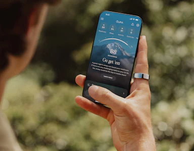 A person views the Oura app on their smartphone while wearing an Oura ring on their right index finger