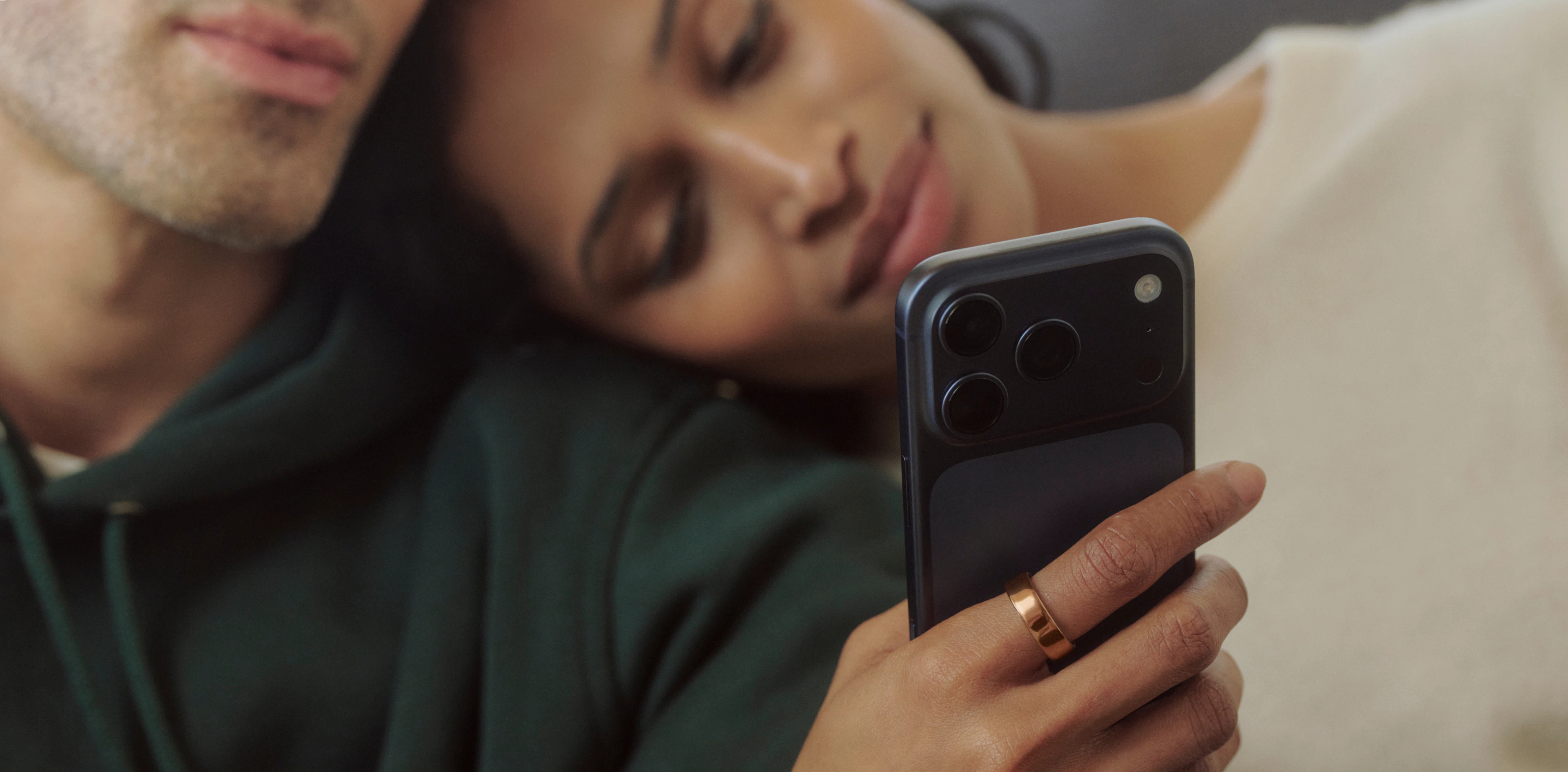 A person wearing a Deep Rose Oura Ring holds a smartphone while resting their head on a companion's shoulder.