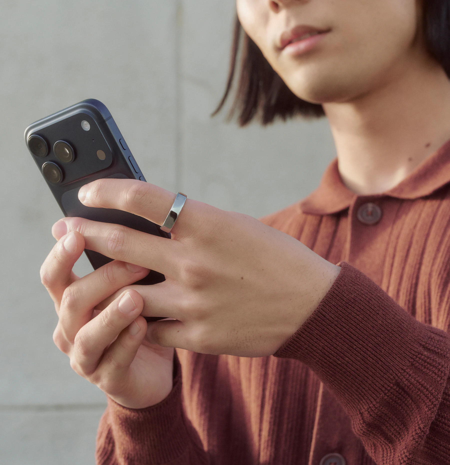 A person holding a smartphone while wearing a Silver Oura Ring on their index finger.