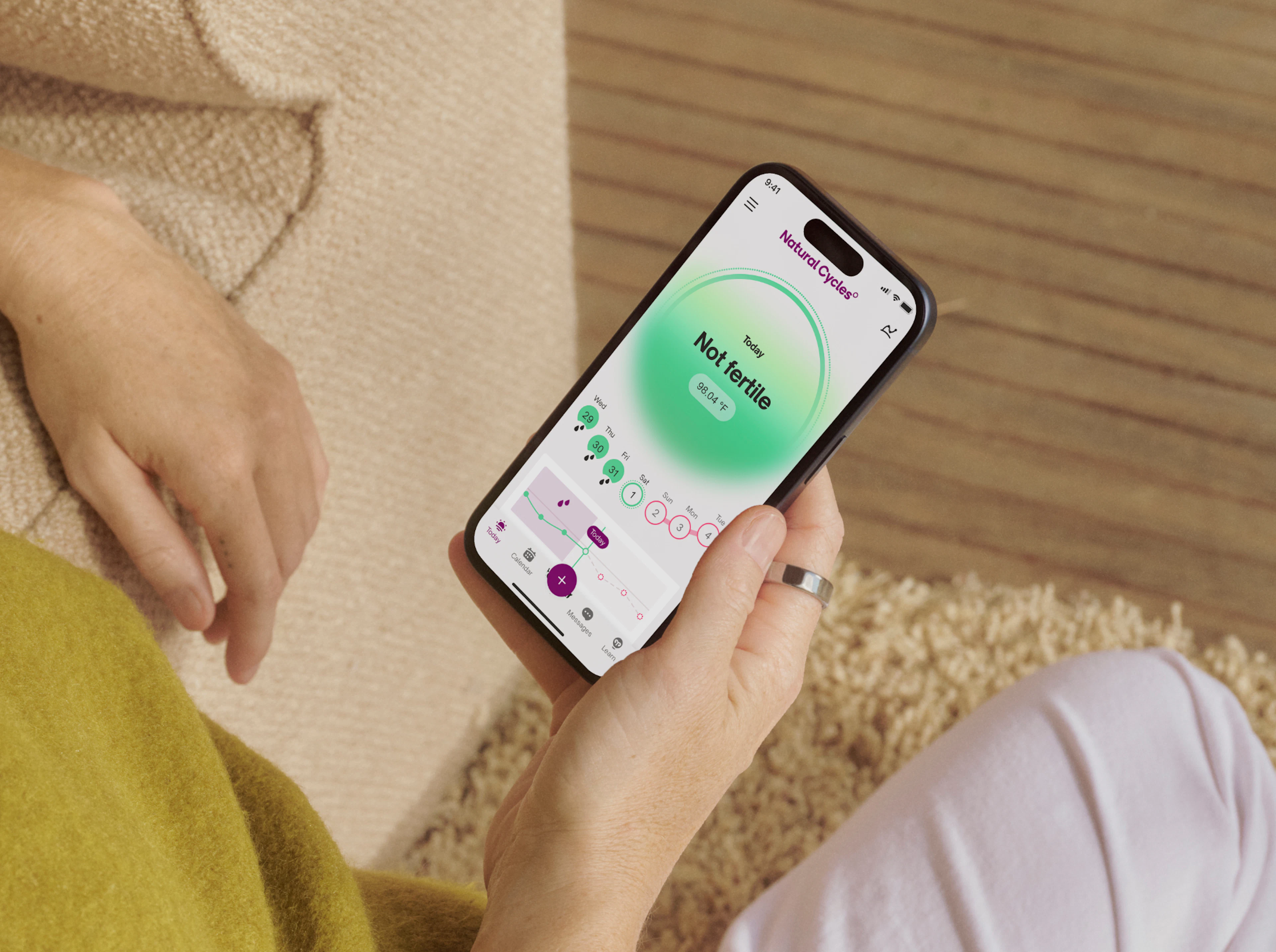 Una persona sujetando un smartphone con la aplicación Natural Cycles abierta en la mano derecha y con un Oura Ring en el dedo índice derecho