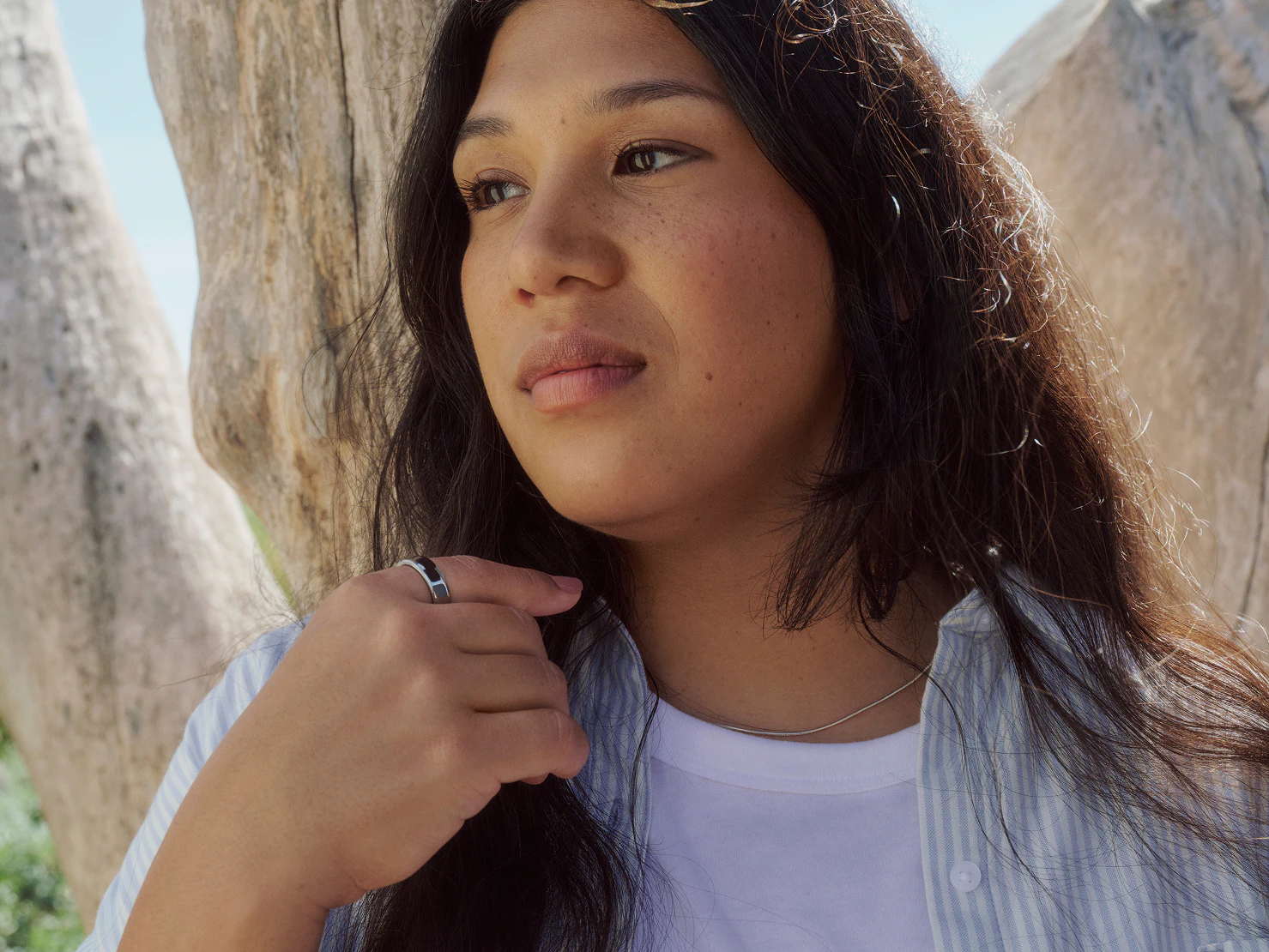 Woman outdoors near sunlit boulders wearing a Silver Oura Ring, hand at her collar.